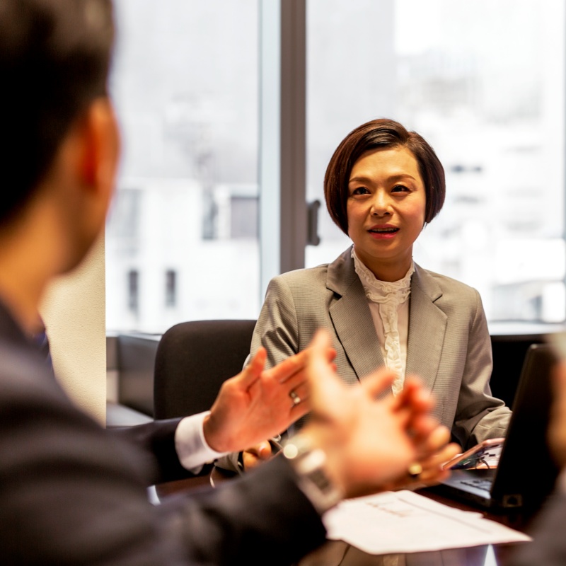 会議室でスーツ姿の女性が、向かいの人物の説明を聞いている様子。