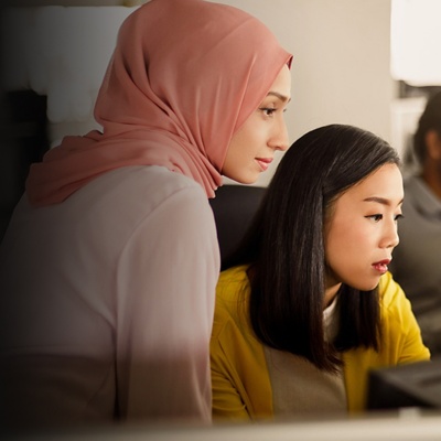 Two women look at an office computer screen while a man talks on the phone in the background.