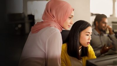 Two women look at an office computer screen while a man talks on the phone in the background.