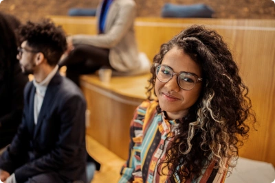 A smiling student with curly hair and glasses sits in a university lecture hall, with other students and tiered seating visible in the background