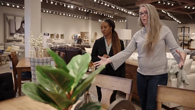 A customer (in grey) and a sales associate (in a black blazer) walking through a furniture showroom.