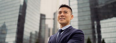 Portrait of a businessman smiling as he looks into the distance, standing with his arms crossed against a background of tall glass covered office buildings in a city