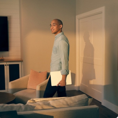 Man holding a laptop, standing in his living room during golden hour. 