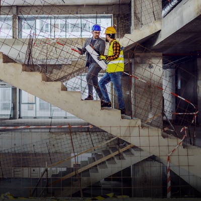 Two men in safety gear discuss plans at a building site