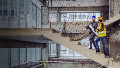 Two men in safety gear discuss plans at a building site