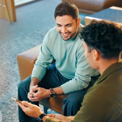 Two male colleagues sitting and chatting in an office lounge area; one male is showing his phone to the other.