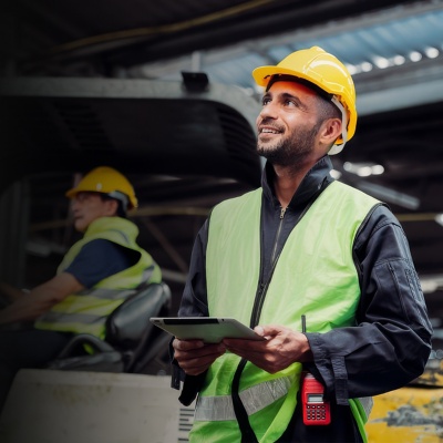 A worker at a construction site holds a tablet and looks beyond the frame