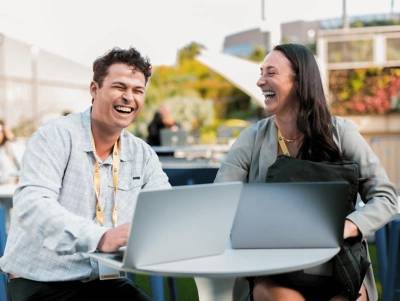 Two people sitting at a table with laptops open and laughing.