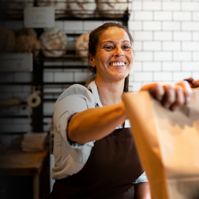 A bakery employee smiles and hands a paper bag to a customer.