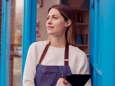 A smiling, professional woman wearing an apron stands confidently in a restaurant or storefront doorway, holding a tablet and looking forward. This image represents a hiring manager or store lead empowered by Conversational ATS technology to go above and beyond and enhance the candidate experience.