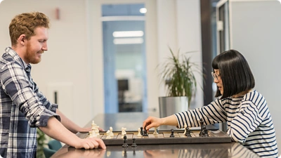 Two employee teammates sitting across from each other, playing chess on the table in between them.