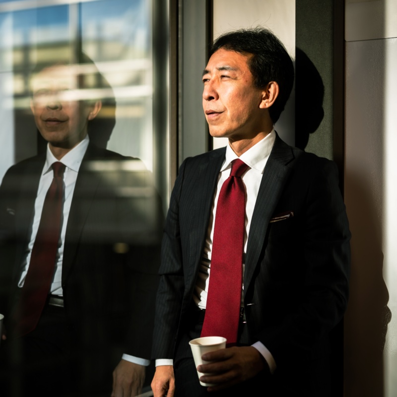 窓辺でコーヒーを持ち、外を見つめる赤いネクタイの男性