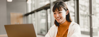 A photo of young woman sitting at a desk in front of her laptop with large windows behind her.