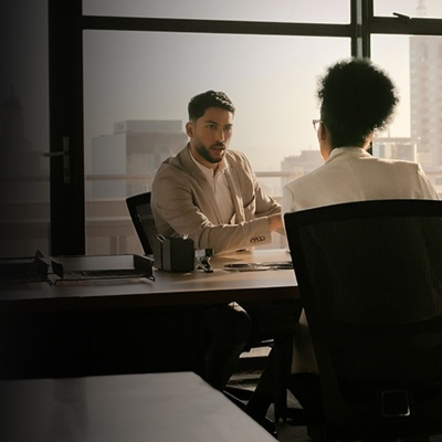 A man sits at a desk in front of a window and speaks to a female colleague seated across from him. 
