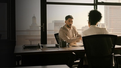 A man sits at a desk in front of a window and speaks to a female colleague seated across from him. 