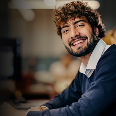 A man with a beard sits at an office desk and smiles at the camera.