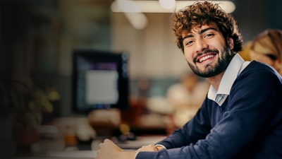 A man with a beard sits at an office desk and smiles at the camera.