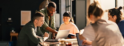 A group of employees in a business casual office sitting around a table looking at a laptop together.