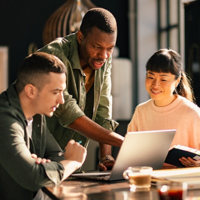 A group of employees in a business casual office sitting around a table looking at a laptop together.