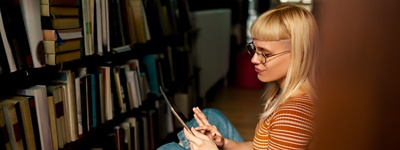 A young student in a library, sitting on the floor, working on an iPad.