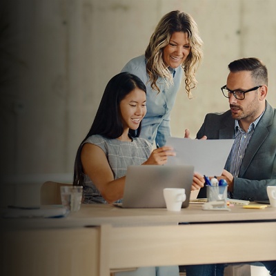 A male and a female coworker sit at a desk looking at a piece of paper while another female employee stands behind them. 