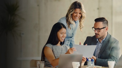 A male and a female coworker sit at a desk looking at a piece of paper while another female employee stands behind them. 