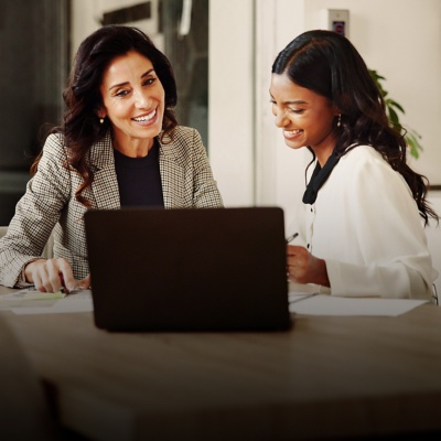Two female colleagues sit at a table and smile while looking at a laptop. 