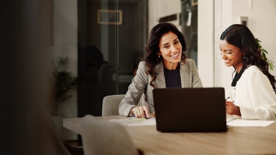 Two female colleagues sit at a table and smile while looking at a laptop. 