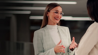 Two women in business attire stand in an office and chat.