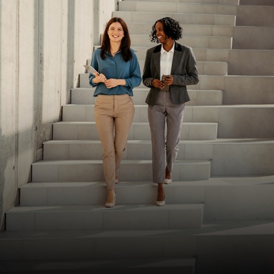 Two female colleagues chat and smile as they descend a staircase. 