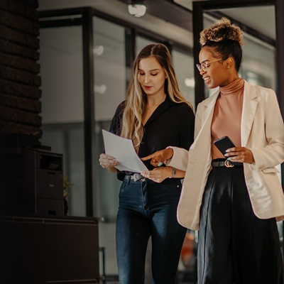 Two female coworkers stand and look down at a document, as one points to something on the page.