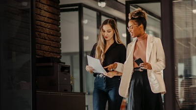 Two female coworkers stand and look down at a document, as one points to something on the page.