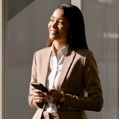 Woman in suit holding phone smiling next to her reflection in the window