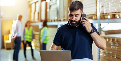 Employee in warehouse working on a laptop and holding a mobile phone