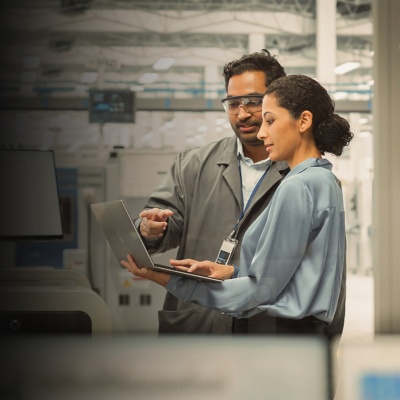 A man and woman discuss data on a laptop screen while standing in a factory