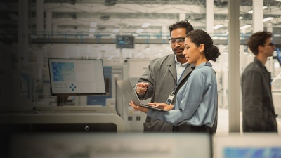 A man and woman discuss data on a laptop screen while standing in a factory